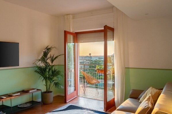 A living room with a sofa, a TV, a potted plant, a sliding glass door leading to a balcony with a chair and a view of the city. - Sorrento Coast Retreat for Families – Terrace & View (Sant'Agnello)