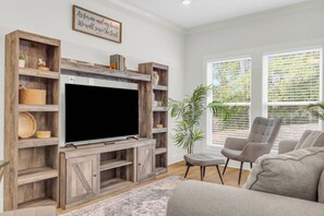 Ein Wohnzimmer mit einem großen Flachbildfernseher, Holzregalen, einem grauen Sofa und einem Blumentopf.