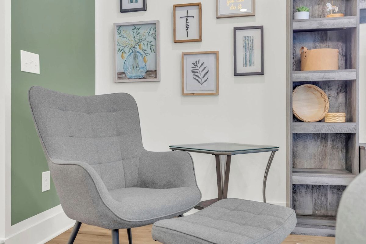 Une pièce comprenant un fauteuil et un pouf recouverts d’un tissu gris, une table avec un plateau en verre et une étagère en bois agrémentée d’objets décoratifs.