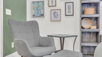 Une pièce comprenant un fauteuil et un pouf recouverts d’un tissu gris, une table avec un plateau en verre et une étagère en bois agrémentée d’objets décoratifs.