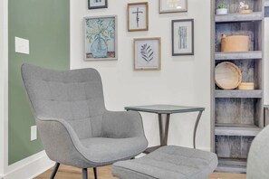 Una habitación con un sillón tapizado gris y un reposapiés, una mesa con superficie de vidrio y un estante de madera con adornos.