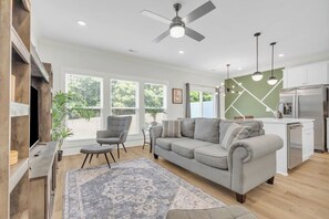 Ein modernes Wohnzimmer mit einem grauen Sofa, einem Couchtisch und einem gemusterten Teppich. Es gibt einen Essbereich mit Tisch und Stühlen sowie eine Küche mit Kühlschrank und einer Kochinsel.