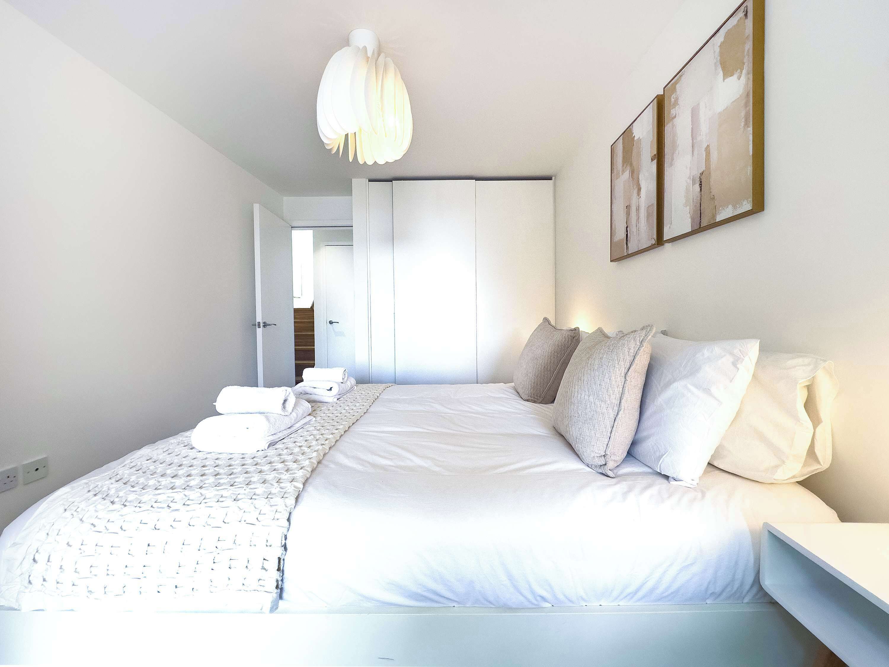 A modern bedroom with a large bed, white bedding, a hanging light fixture, and a painting on the wall.