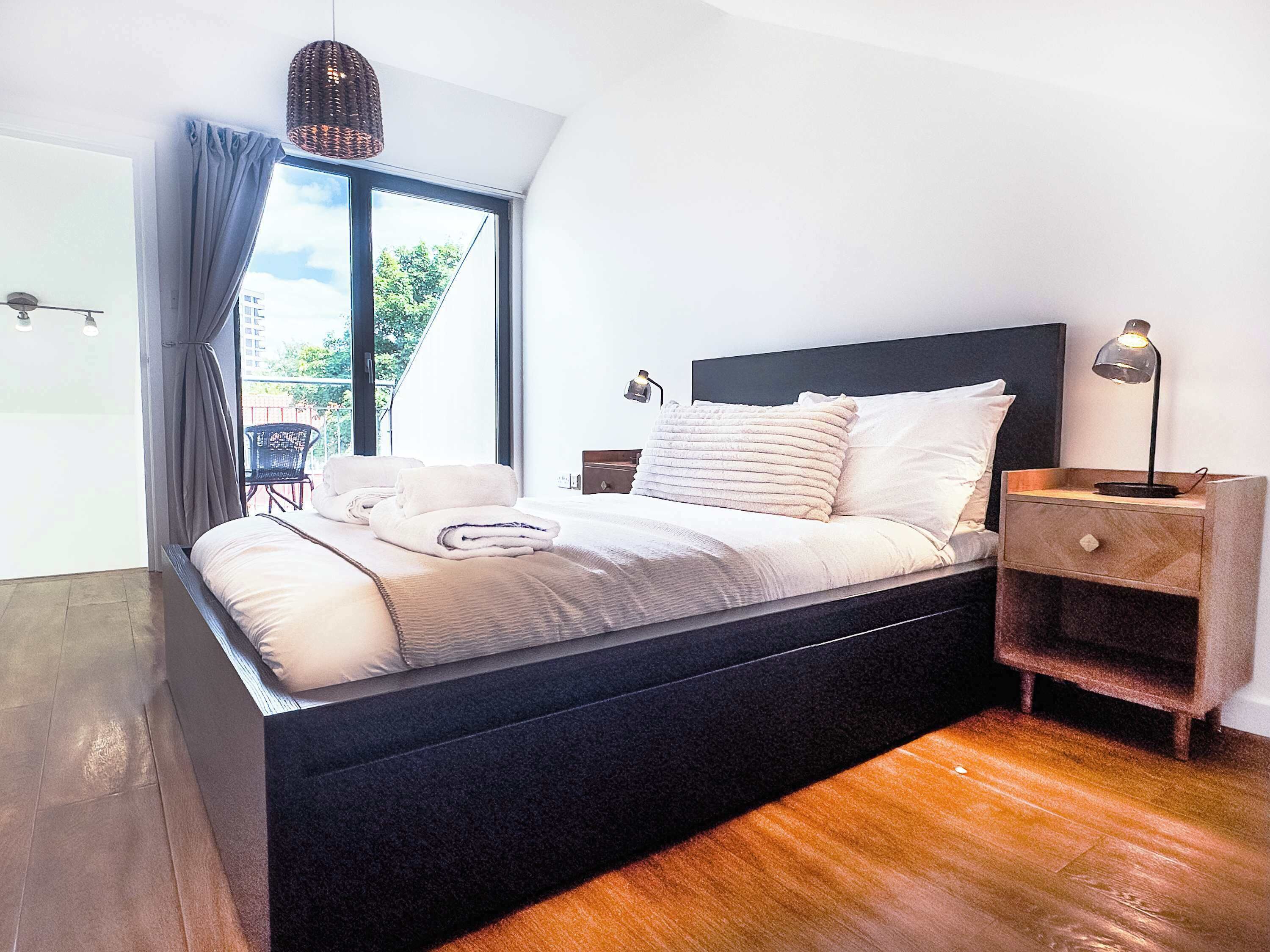 A modern bedroom with a large bed, a nightstand, a lamp, and a view of the outdoors through a window.