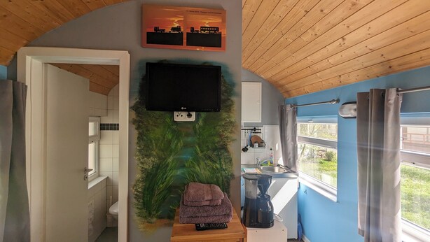 A compact living space with a flat-screen TV mounted on the wall, a small table with a remote control, and a window with curtains. - Spend the night in a railroad carriage - nostalgia & comfort in Thuringia (Kirchheilingen)