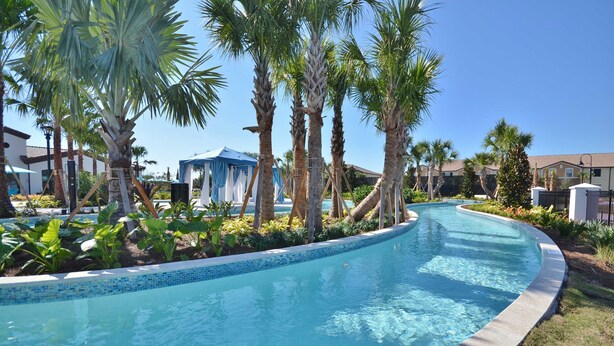 A swimming pool with a tiled edge, surrounded by palm trees and greenery, with buildings in the background.