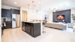 A modern kitchen with a central island, stainless steel appliances, pendant lighting, and a living area with a sofa and TV.