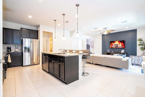 A modern kitchen with a central island, stainless steel appliances, pendant lighting, and a living area with a sofa and TV.