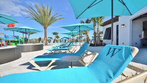 A poolside area with blue lounge chairs and umbrellas, palm trees, and a children's play area in the background.