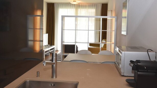 Een compacte hotelkamer met een kitchenette, een zithoek met tafel en stoelen, een groot raam met gordijnen, en een bed op de achtergrond.