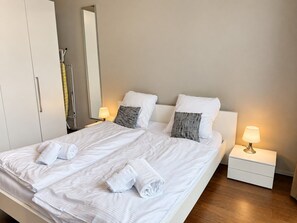 Een tweepersoonsbed met witte lakens en twee kussens, een nachtkastje met een lamp, en een garderobe.