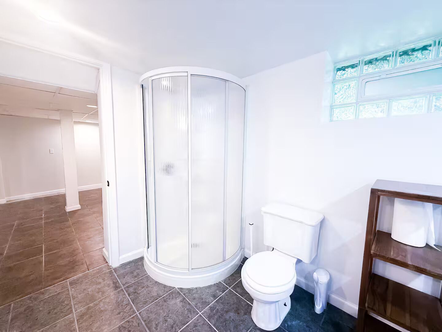 A bathroom with a glass shower enclosure, a white toilet, and a wooden vanity with shelves.