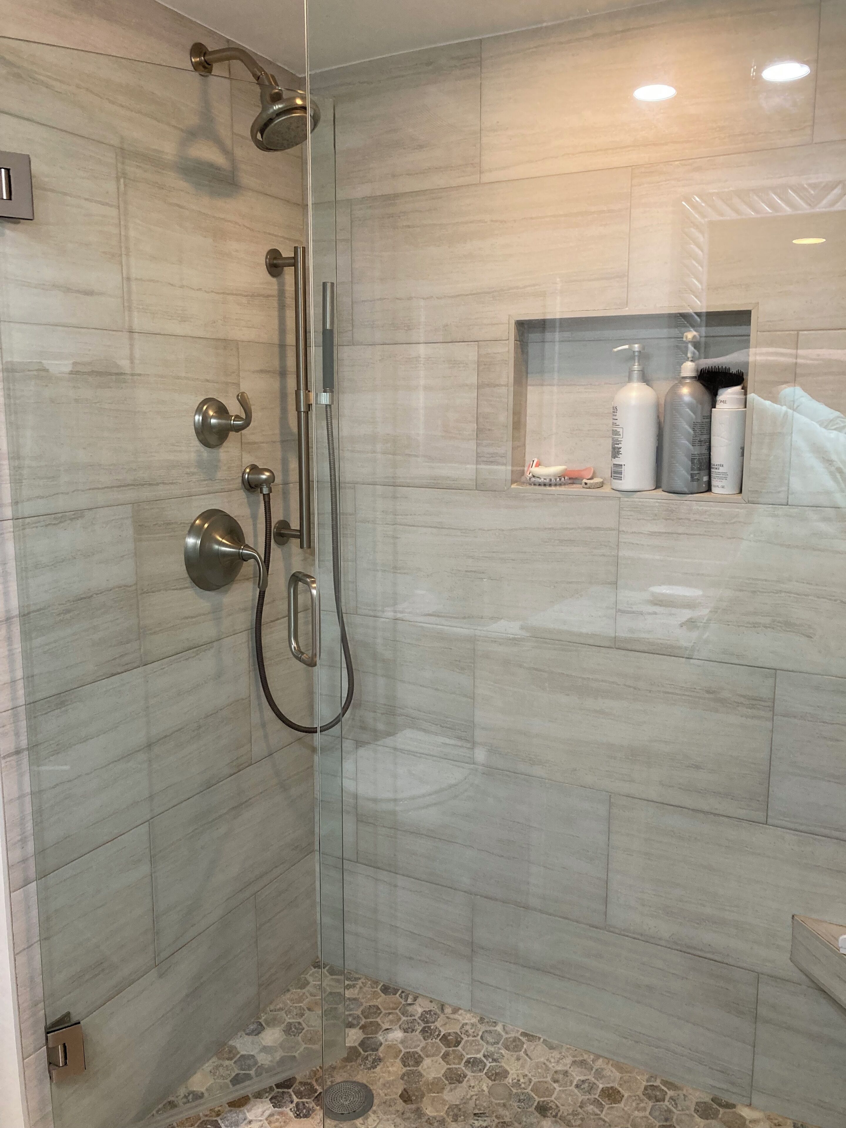 Eine moderne Dusche mit Glasabtrennung, Handbrause, Wandarmatur und Ablagefläche für Toilettenartikel.