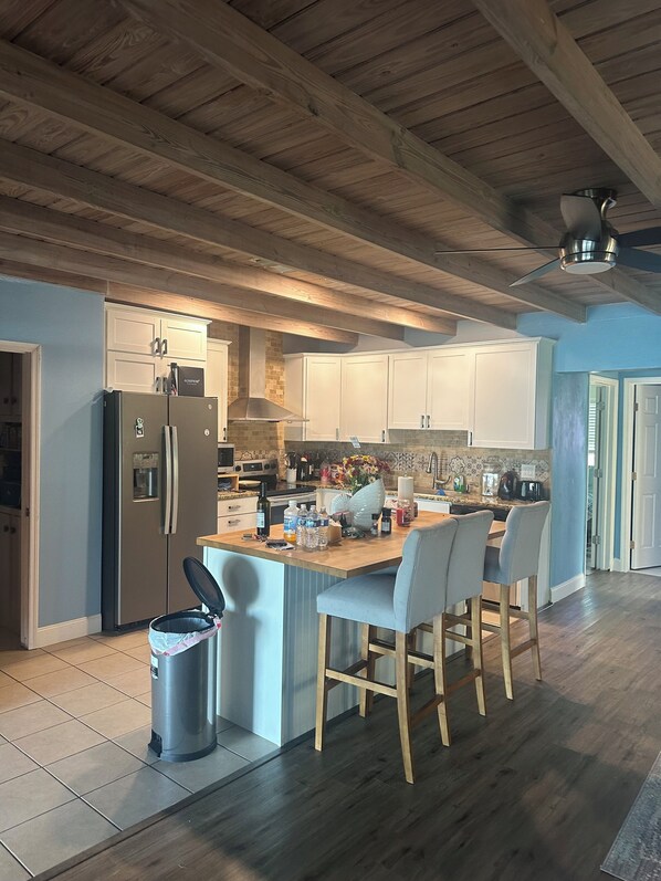 A kitchen with wooden beams, a central island with chairs, a refrigerator, and a trash can.