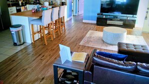 Una sala de estar con un sofá, un reposapiés y una mesa con un libro. Hay un televisor, una cocina al fondo y un bote de basura.