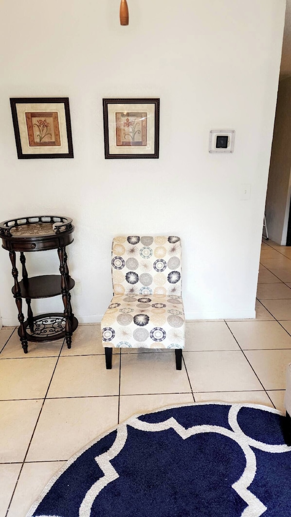 A room with a patterned armchair, a small round table, two framed pictures on the wall, and a blue rug on the floor.