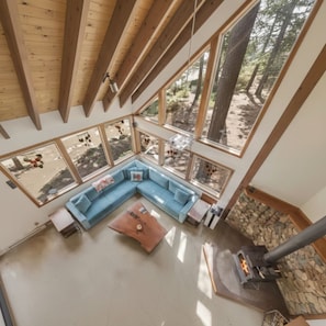 A high-ceilinged room with a large window overlooking a forest, a stone fireplace, and a blue sectional sofa. - Tahoe Summer Escape • Theater, Gym & Next to Lake! (Tahoe City)