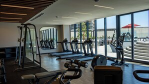 Une salle de sport moderne équipée de tapis de course, d’appareils de musculation et d’un banc. De grandes fenêtres offrent une vue sur l’extérieur.