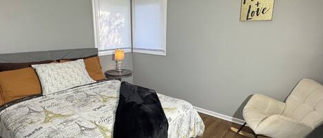 A bedroom with a bed, a rocking chair, a nightstand with a lamp, and a window with blinds.