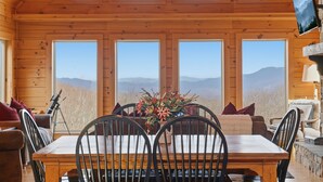 A wooden cabin interior with a dining table, chairs, and a large window offering a mountain view.
