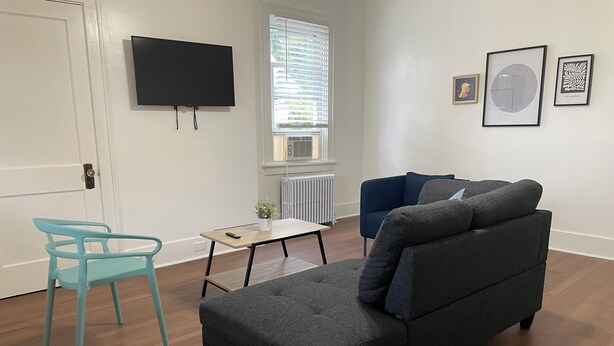 Una sala de estar con un sofá gris, una silla azul, una mesita, un televisor y obras de arte enmarcadas en la pared.