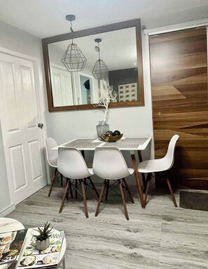 Un coin repas avec une table blanche, quatre chaises blanches, un plancher en bois, un miroir au-dessus de la table et une porte en bois sur la droite.