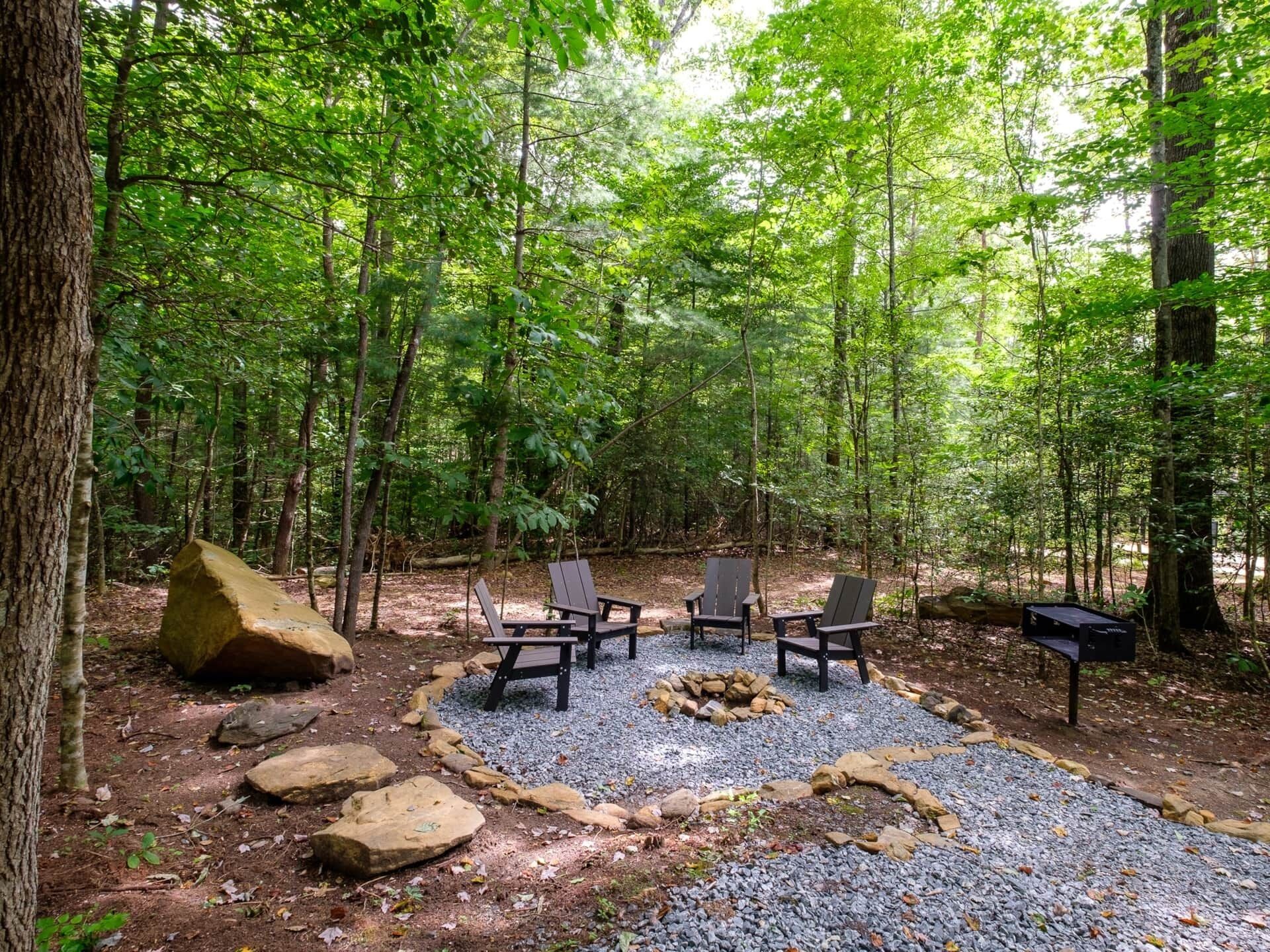 A forest campsite with a fire pit, four Adirondack chairs, and a mailbox.