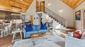 A spacious living area with a high ceiling, wooden beams, a staircase, and a kitchen with modern appliances. - Last Minute Deals! ~Fire Pit, Hot Tub,  on Creek! (Vilas)