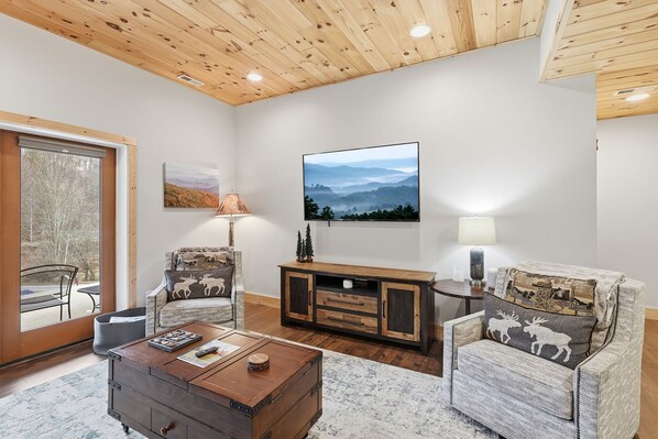 A cozy living room with a wooden ceiling, a large window, a sofa, a coffee table, and a cabinet with a landscape painting.