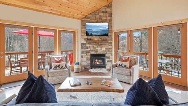 A cozy living room with a stone fireplace, comfortable seating, and large windows offering scenic views. - Last Minute Deals! ~Fire Pit, Hot Tub,  on Creek! (Vilas)