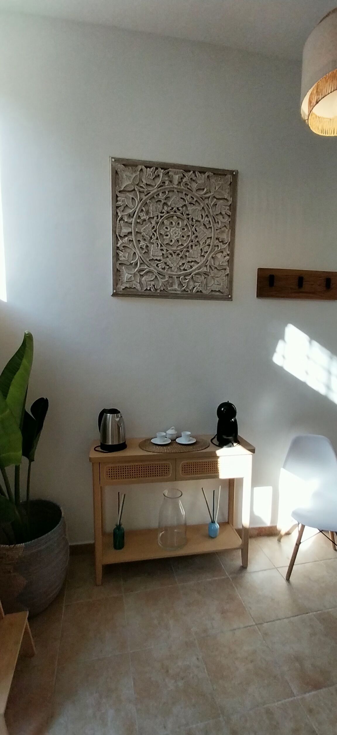 A room with a wooden side table, a decorative wall piece, a potted plant, a coffee maker, and a white chair.