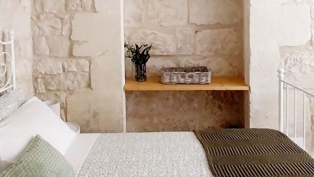 A bedroom with a stone wall, a bed with patterned bedding, a hanging light fixture, and a shelf with a plant and basket.