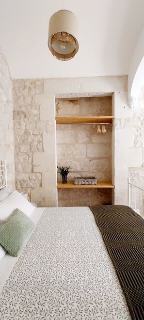 Quarto com parede de pedra, cama com roupa de cama estampada, luminária pendente e prateleira com planta e cesta.