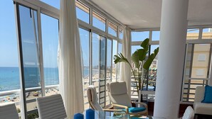 A modern living room with a glass table, white cushioned chairs, and a view of the beach.