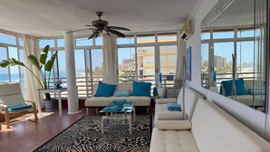A modern living room with a sofa, a chair, a coffee table, and a large window offering a view of the beach and cityscape.