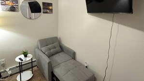 Una habitación compacta con un sofá gris, un televisor montado, un espejo redondo pequeño y una mesita de noche con una planta.