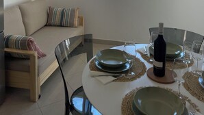 Een eetgedeelte met een ronde tafel gedekt voor twee personen, een rieten hanglamp, uitzicht op de zee door een raam, en een bank met gestreepte kussens.