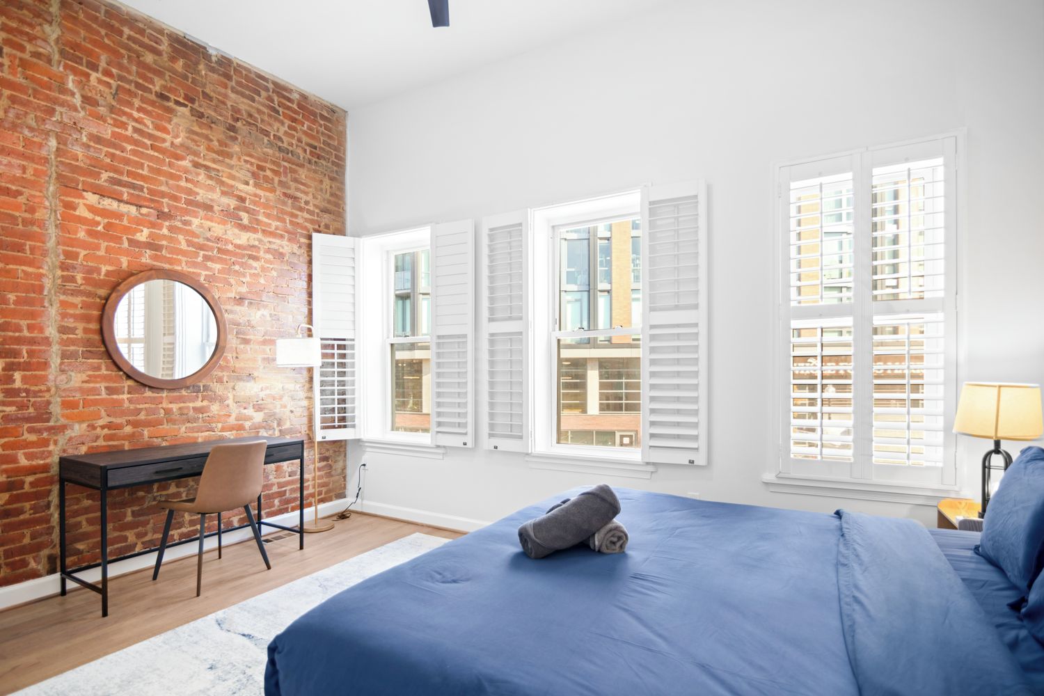 Un dormitorio con pared de ladrillo, un escritorio, una silla, una cama con ropa de cama azul y ventanas con contraventanas.