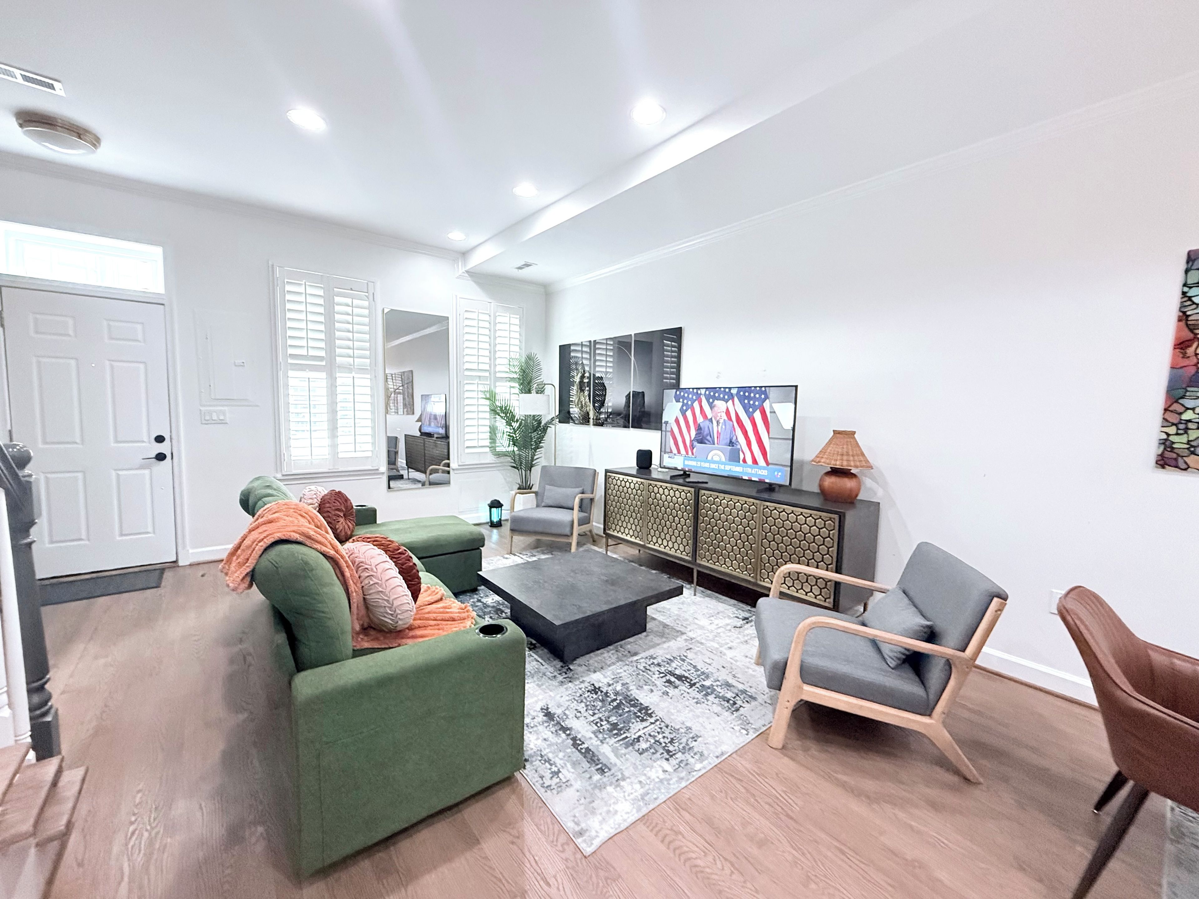 Un salon moderne avec un canapé vert, un fauteuil gris, un pouf noir et un téléviseur affichant un drapeau américain en arrière-plan.