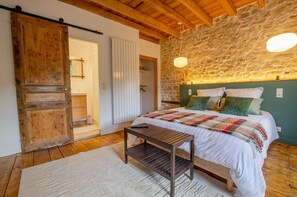 A bedroom with a stone wall, wooden ceiling beams, a bed with multiple pillows, a bedside table, and a sliding door. - Le Clos de la Ferme - CHERVEY (CHERVEY)