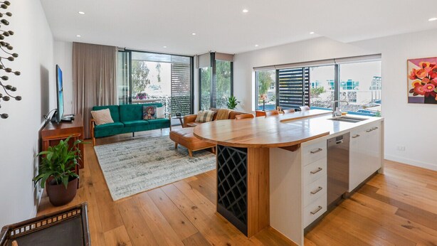 A modern kitchen with a central island, large windows, and a green sofa in the living area.