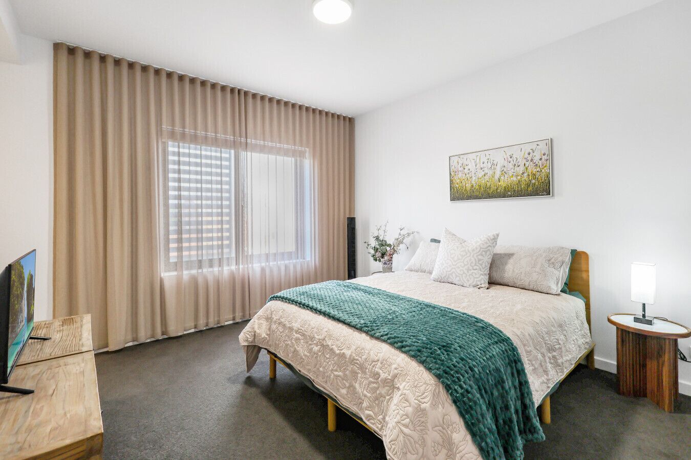 Ett hotellrum med en säng, ett sidobord, en lampa, en tavla och ett fönster med gardiner.