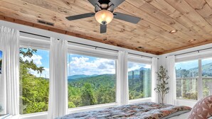 Un dormitorio con una cama grande, un ventilador de techo y varios ventanales con vistas a un paisaje boscoso.