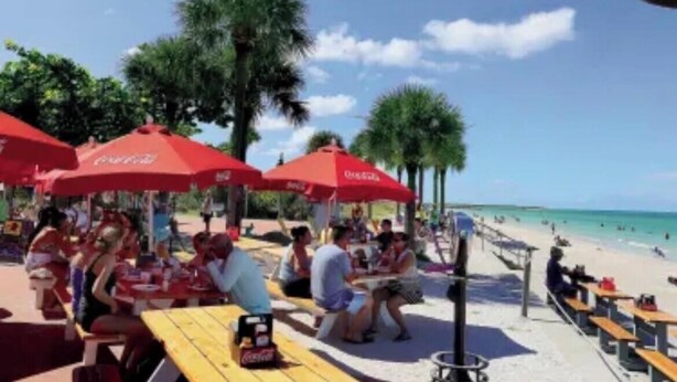 A beachside dining area with red umbrellas and wooden tables.