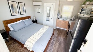 A bedroom with a bed, bedside table, and a small nightstand. There is a door leading to a bathroom and a kitchenette area with a refrigerator and a sink. - Olde Town Arvada Tiny Retreat (Arvada)