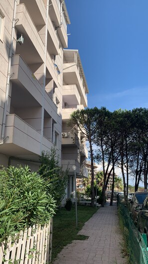 A multi-story residential building with balconies, a paved pathway, and a row of trees. - One Bedroom Apartment  fully furnished with AC in charming Qerret Beach, Albania (Qeret)