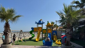 A playground with slides and climbing structures, surrounded by palm trees and beach chairs. - One Bedroom Apartment  fully furnished with AC in charming Qerret Beach, Albania (Qeret)