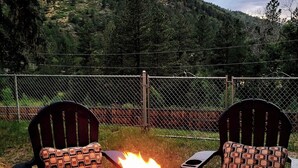 A fire pit with burning logs, surrounded by two chairs with patterned cushions, set against a backdrop of a forested hillside. - Mountain Escape Near RMNP, Views, River Access, Dogs OK (Drake)