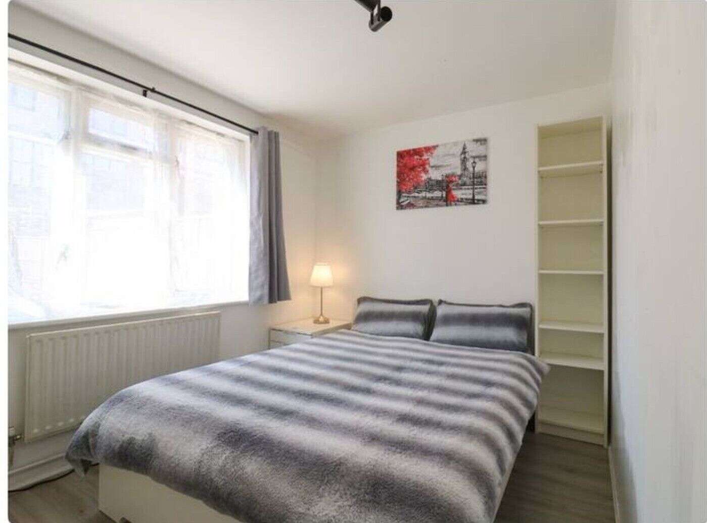 Une chambre à coucher avec un lit, une fenêtre avec des rideaux, une lampe de chevet, une étagère fixée au mur et un tableau.