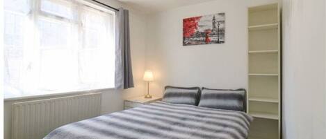 Une chambre à coucher avec un lit, une fenêtre avec des rideaux, une lampe de chevet, une étagère fixée au mur et un tableau.